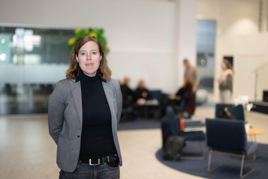 En kvinna i grå kavaj och svart tröja står i förgrunden i en ljus och modern kontorsmiljö. I bakgrunden syns sittplatser och flera personer.