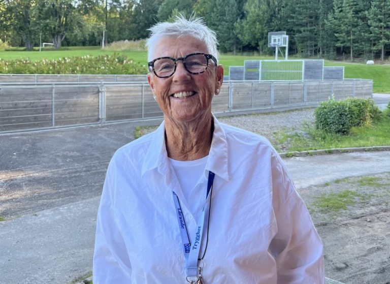 ”En äldre person med kort, vitt hår och mörka glasögon står utomhus och ler mot kameran. Personen är klädd i en vit skjorta och bär ett blått nyckelband med nycklar. I bakgrunden syns en grön trädgård med träd, buskar och en liten basketkorg samt ett staket.”