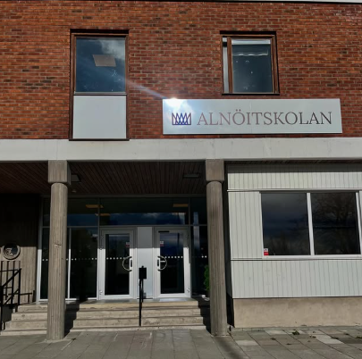 : Entrén till Alnöitskolan, en tegelbyggnad med en skylt med skolans namn och logotyp ovanför de dubbla glasdörrarna, samt en ramp och trappa fram till ingången.