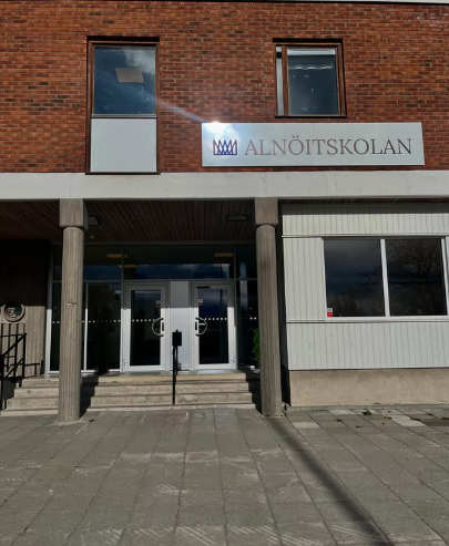 : Entrén till Alnöitskolan, en tegelbyggnad med en skylt med skolans namn och logotyp ovanför de dubbla glasdörrarna, samt en ramp och trappa fram till ingången.