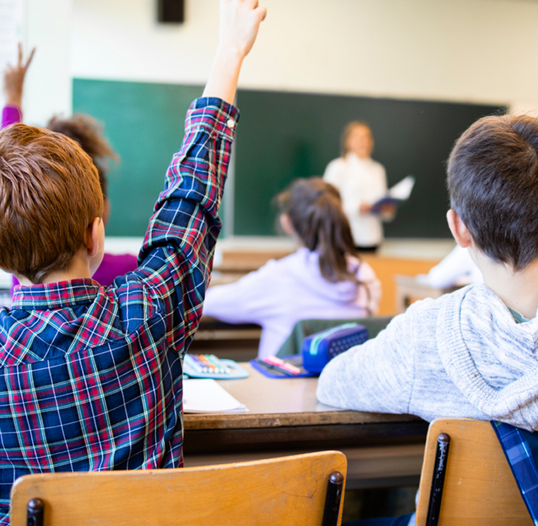 En elev räcker upp handen i ett klassrum, sedd bakifrån, medan läraren står längst fram vid tavlan med en bok i handen. Andra elever sitter vid sina bänkar och följer undervisningen.