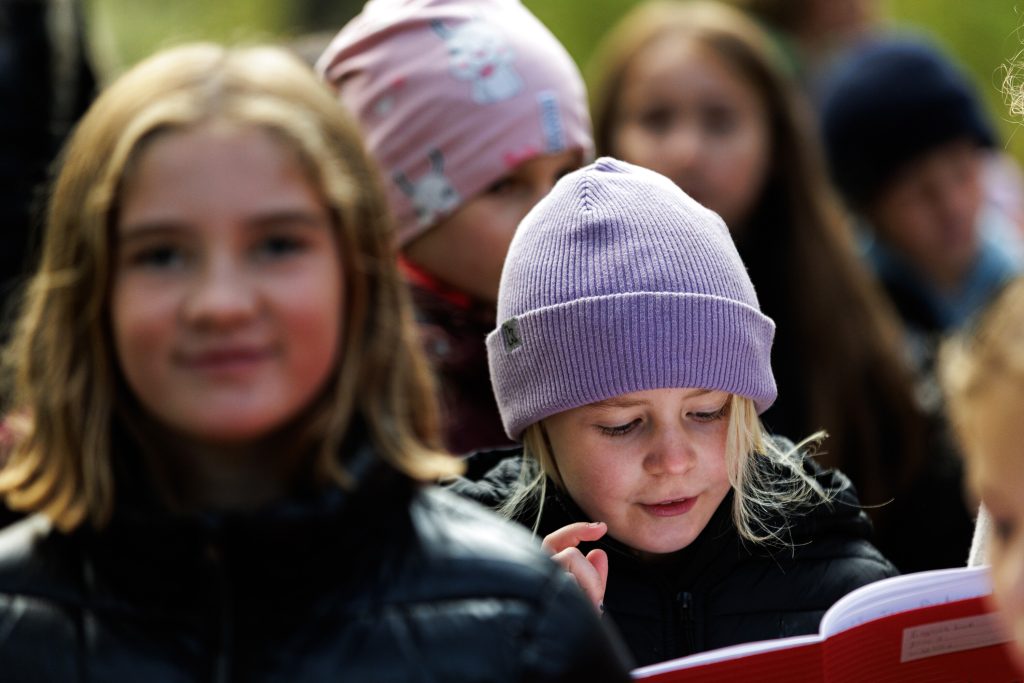 : Ett barn i lila mössa tittar koncentrerat ner i en röd bok utomhus, omgiven av andra barn i mössor och jackor som syns suddigt i förgrunden och bakgrunden.
