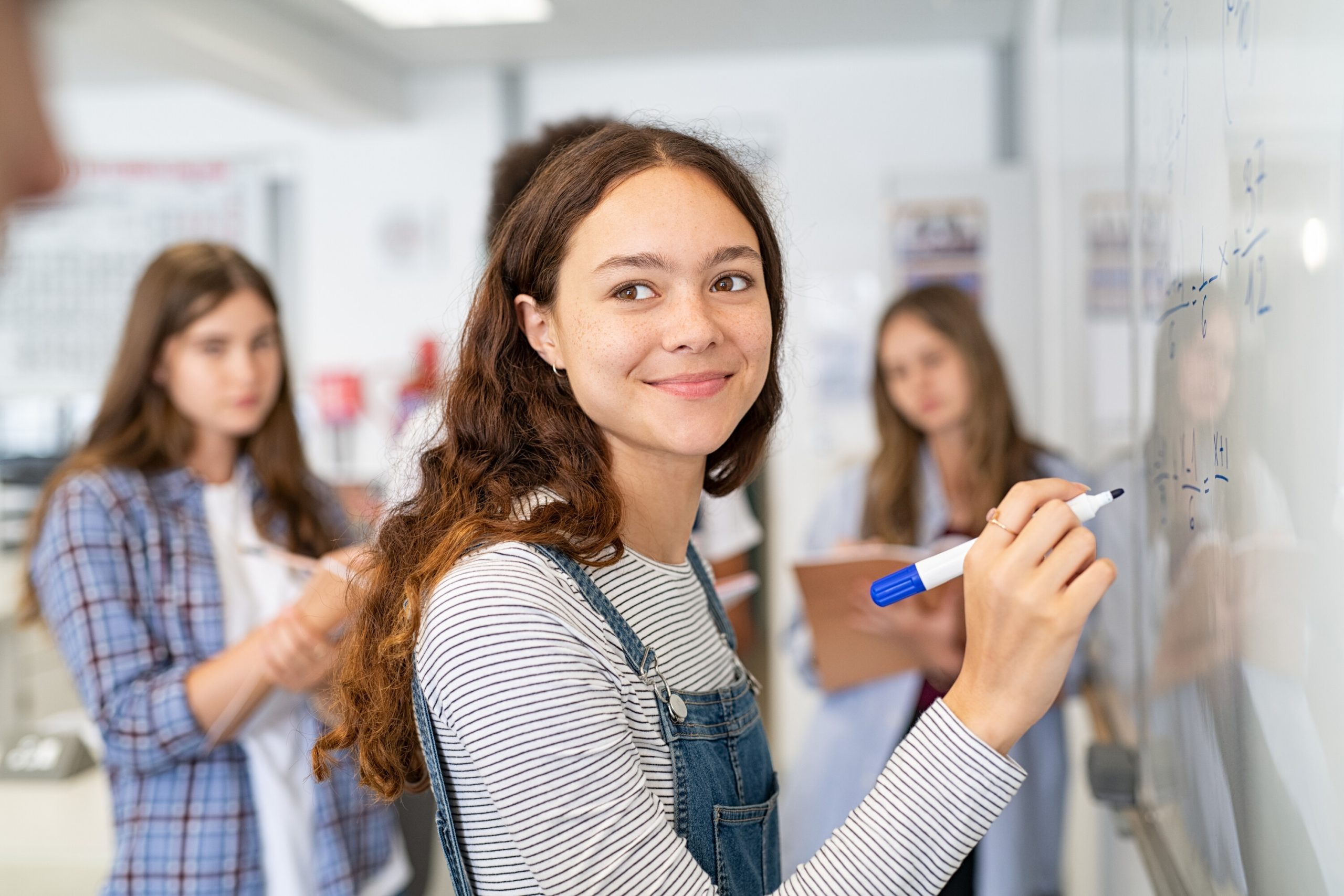 En leende elev i randig tröja och jeansoverall står vid en whiteboardtavla med matematiska ekvationer och håller en blå whiteboardpenna, medan hon tittar in i kameran. I bakgrunden syns klasskamrater som arbetar i klassrummet.