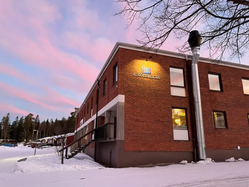 Alnöitskolans tegelbyggnad fotograferad i vinterlandskap med snötäckt mark, under en himmel färgad i rosa och blått vid skymning.