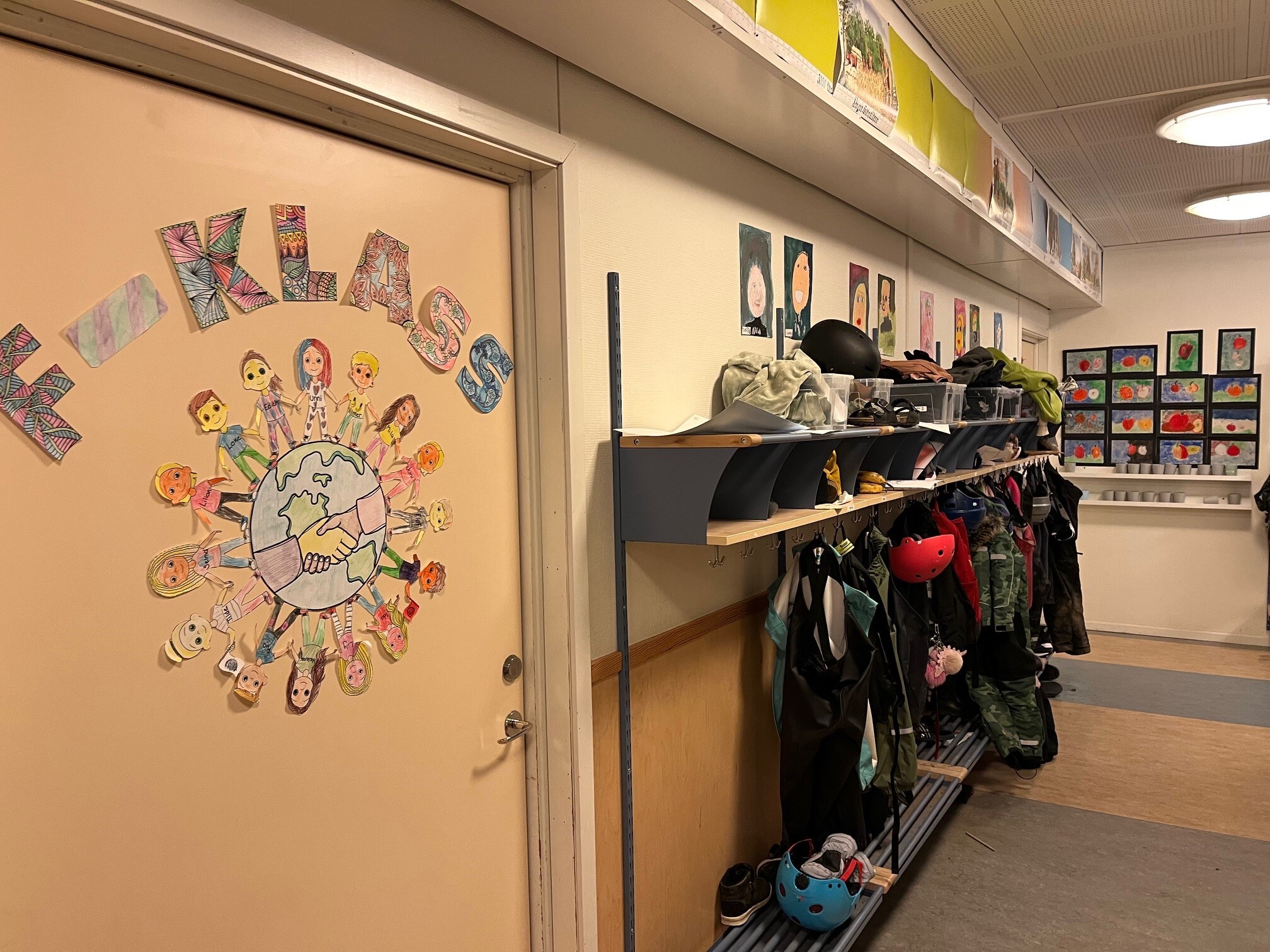 En skolkorridor med en dörr dekorerad med elevernas handgjorda figurer runt ett jordklot och texten "F-klass". Bredvid dörren syns en klädhylla med jackor, hjälmar och skor, samt elevernas teckningar uppsatta på väggen längs korridoren.