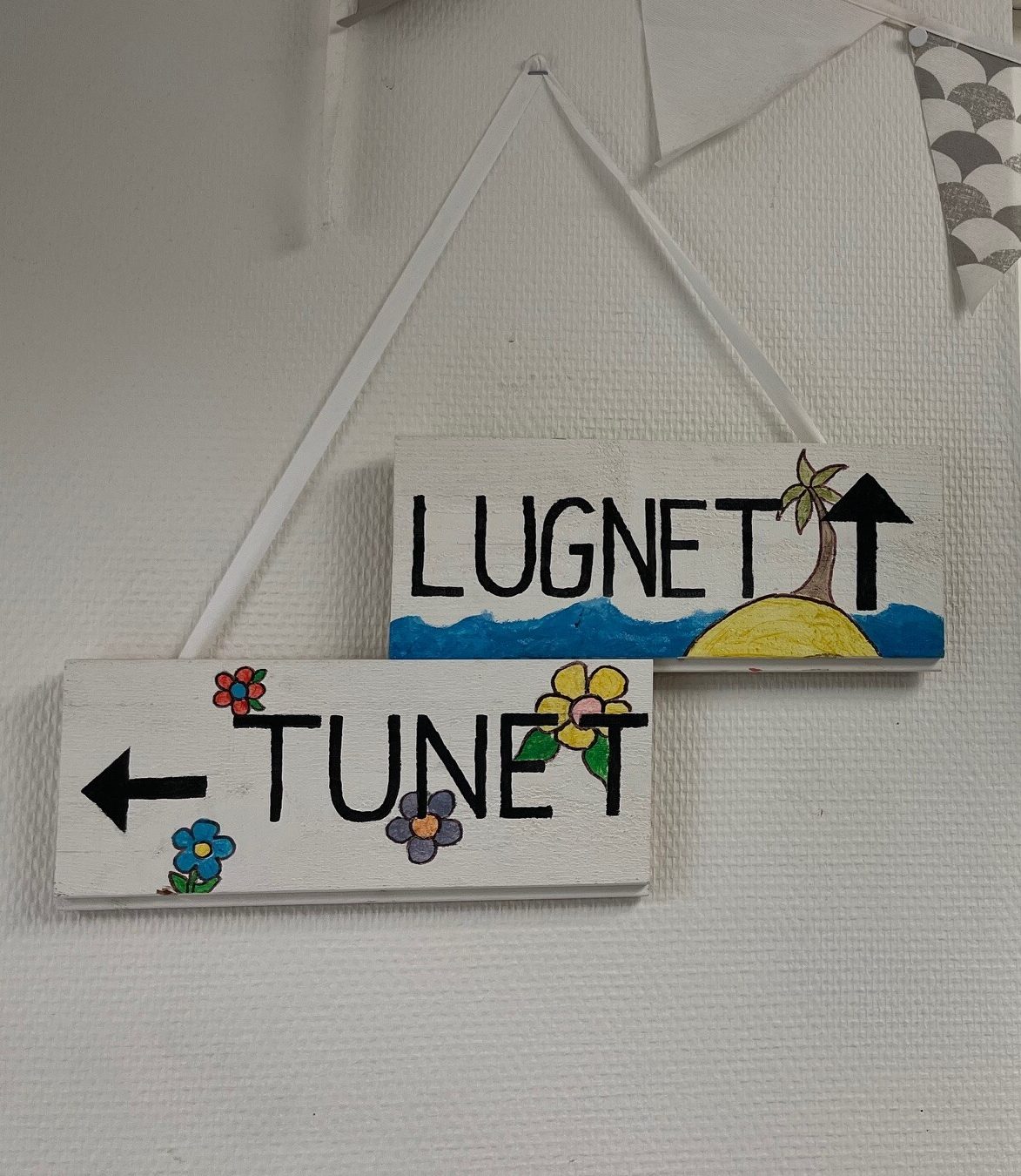Två handmålade träskyltar hänger på en vägg. Den ena visar texten "Tunet" med en pil åt vänster och målade blommor, den andra visar texten "Lugnet" med en målad ö, palm och hav.