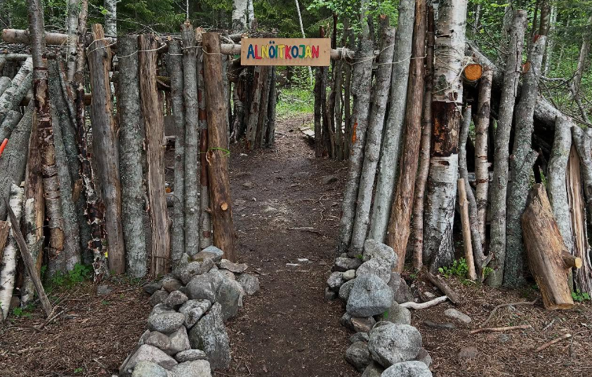 En stig kantad av upprätta trädstammar och grenar leder genom en skog till en skylt med texten "ALNÖITKOJAN", omgiven av grönska och stenar längs marken.