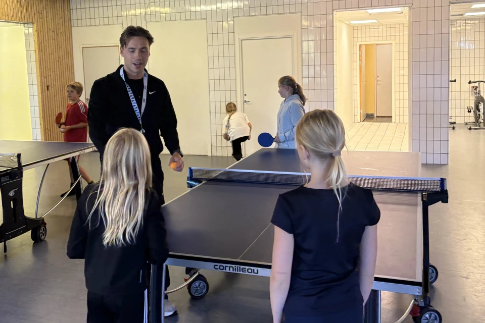 En vuxen ledare i svart träningsjacka och med nyckelband runt halsen står vid ett bordtennisbord och håller en orange pingisboll medan han pratar med två blonda flickor som står med ryggen mot kameran. Rummet är en ljus lokal med kakelväggar och dörrar. I bakgrunden spelar fler barn pingis vid ett annat bord, och en pojke i röd tröja håller ett racket. Genom en öppning syns ett angränsande rum med en träningscykel.